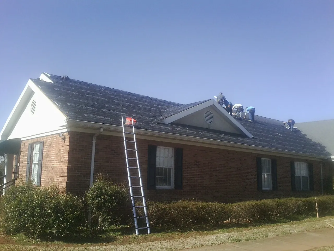 Crew tearing shingles off a brick ranch roof with a ladder set — Metal Roof Repair work by our crew in Lake Station
