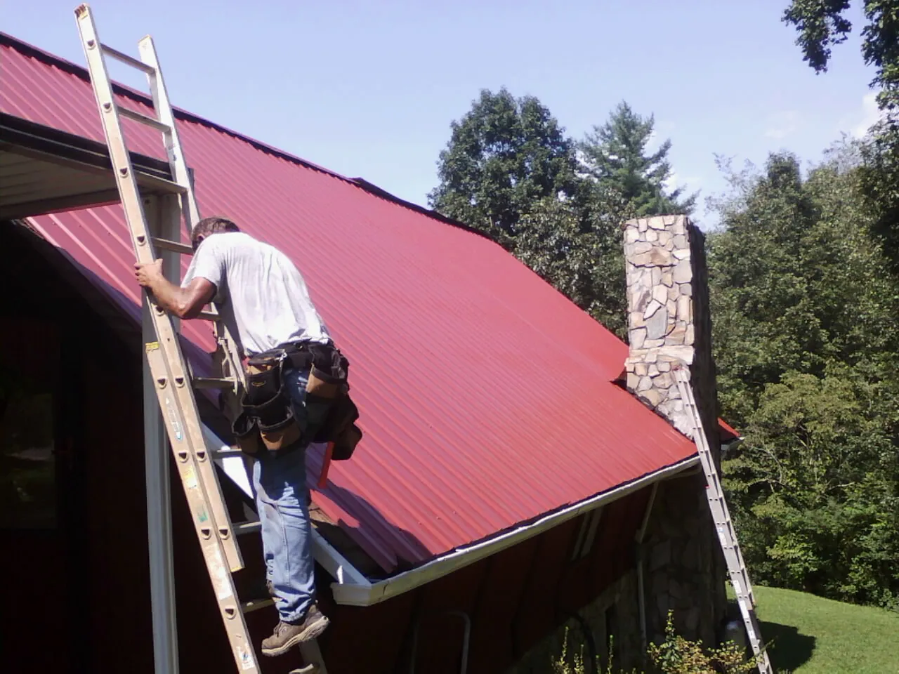 Roofer on a ladder beside a red metal roof and stone chimney — Soffit & Fascia Repair by Roofing Brothers in Lake Station